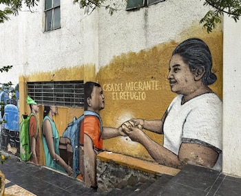 Mural en pared blanca y amarilla, con una mujer entregando un objeto a un joven con mochila. Otros dos jóvenes con mochilas y la inscripción "Casa del Migrante El Refugio"