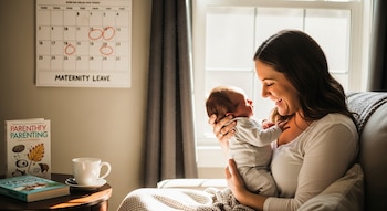 Mujer sonriente acuna a un bebé recién nacido en un sofá, con un calendario de "MATERNITY LEAVE" y fechas circuladas visible en la pared trasera.