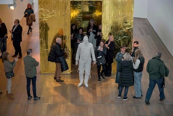 Una persona con un disfraz de momia blanca está de pie en una galería de museo, rodeada de gente diversa que toma fotos, con una pared dorada arrugada al fondo