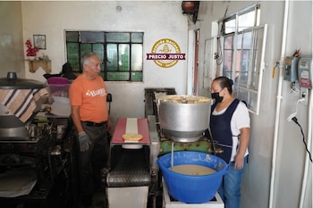 Hombre y mujer en tortillería. La mujer opera máquina que procesa masa de maíz, vertiéndola en un cubo azul, con una tortilla de maíz fresca en la cinta transportadora