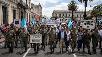 Vista de veteranos de guerra de Guatemala marchando en la calle, llevando uniformes de camuflaje y bastones. Portan pancartas y banderas de Guatemala. Edificios y palmeras visibles en el fondo.