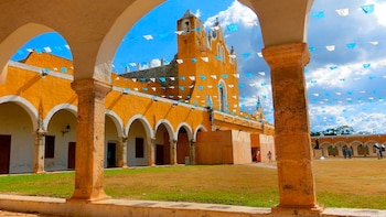 Arcos de piedra de color amarillo rodean un patio con banderines azules y blancos colgados frente a la fachada de una iglesia colonial
