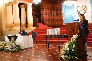 Seis personas formadas en fila sostienen folletos impresos en un salón frente a la bandera de Guatemala y arreglos florales