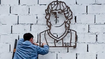 Hombre instala una figura metálica de busto en una pared blanca de ladrillos. La figura representa a Diego Maradona y tiene el número 10