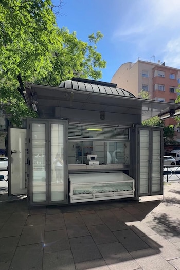 Un moderno kiosco gris con puertas de vidrio y un mostrador con una vitrina abierta, ubicado en una acera arbolada bajo un cielo azul brillante en Madrid