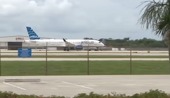 Avión blanco de JetBlue con cola azul y el logo de la aerolínea en el fuselaje, en un aeropuerto con hangares, césped, y una valla metálica