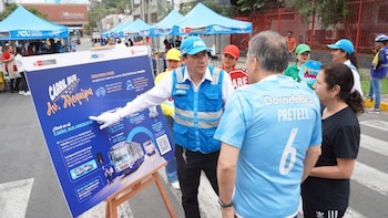 Un trabajador con chaleco azul señala un cartel informativo sobre el carril de buses ante dos personas en una calle con carpas azules