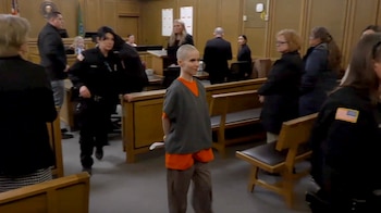 Alyssa Bradburn, con la cabeza rapada y vestimenta de prisión, camina por una sala de tribunal con bancos de madera, custodiada por oficiales