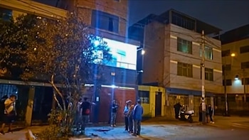 Vista nocturna de una calle con edificios residenciales, personas congregadas, un árbol frondoso, un poste de luz y una motocicleta estacionada