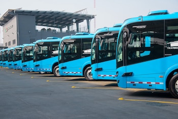 Vista lateral de una fila de autobuses azules marca Scania estacionados en un patio, con un edificio gris al fondo bajo un cielo claro