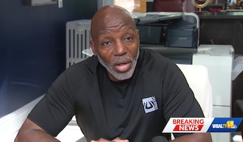 Un hombre calvo, de mediana edad y barba gris, con camiseta negra, habla en un entorno de oficina. Un rótulo de 'BREAKING NEWS' es visible