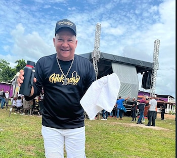 Hombre sonriente, vestido con camiseta negra, pantalones blancos y gorra, sostiene una botella negra y una toalla blanca frente a un escenario al aire libre con personas