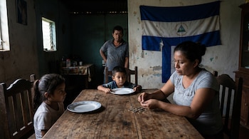 Una mujer sentada en una mesa de madera cuenta monedas, con dos niños de platos vacíos a su lado. Un hombre preocupado de pie al fondo, con una bandera de Nicaragua en la pared.