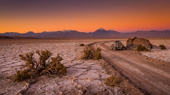 Un vehículo 4x4 oscuro en un camino de tierra en un desierto salino. Se ven montañas nevadas, un cielo de atardecer naranja y plantas secas.