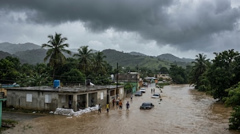 Vista aérea de una calle rural en República Dominicana inundada con agua marrón, personas caminan y autos están sumergidos, con edificios y vegetación al fondo bajo un cielo nublado.