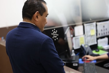 Un hombre de perfil con saco azul ante un mostrador con mampara de vidrio, un monitor y un teclado de computadora