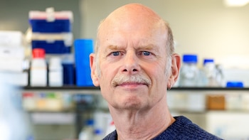 Retrato de un hombre calvo con bigote claro y ojos azules, vistiendo un suéter azul oscuro, en un laboratorio con estantes y material científico borroso detrás