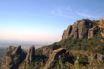 Un paisaje natural con montañas rocosas, vegetación densa y árboles. El cielo es azul claro con nubes finas. Al fondo se observa una ciudad con bruma