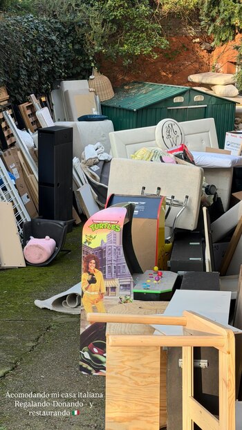 Gran pila de muebles, como cabeceros de cama y sofás, cajas y objetos varios acumulados en un patio de cemento con musgo. Al fondo, un cobertizo verde y vegetación frondosa