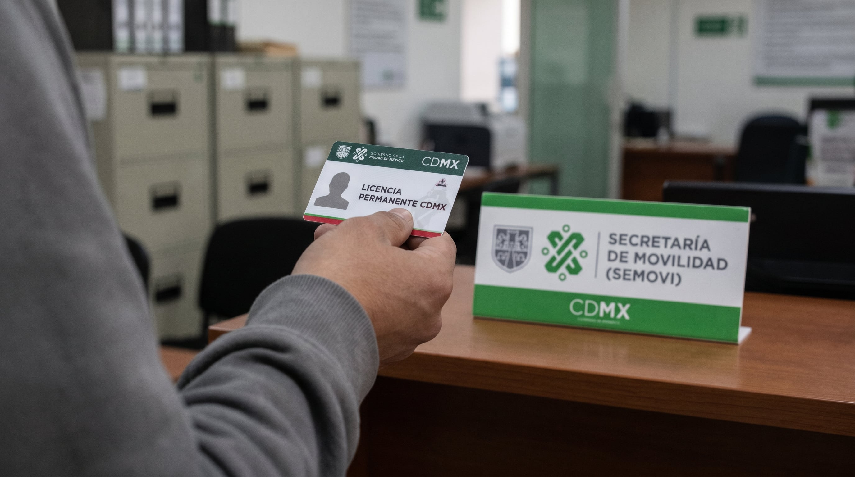 Una mano sostiene una Licencia Permanente de Conducir de la Ciudad de México en una oficina gubernamental, con el letrero de la Secretaría de Movilidad (Semovi) en el fondo. (Imagen Ilustrativa Infobae)