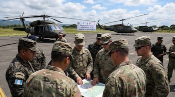 Soldados de Guatemala y EE.UU. revisan un mapa en una pista, con helicópteros militares y una pancarta de "Operación Conjunta" contra narcotráfico.