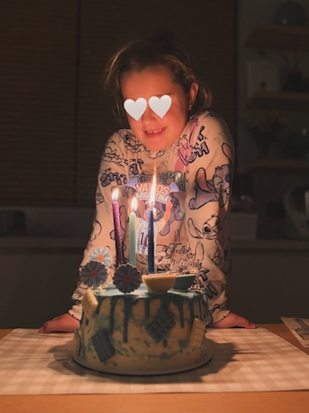Una niña sonriente, con ojos cubiertos por emojis de corazón, mira un pastel de cumpleaños azul con cuatro velas encendidas sobre una mesa. Fondo oscuro