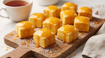 Porciones cuadradas de bizcocho con cobertura de naranja sobre una tabla de madera espolvoreada con azúcar y una taza de té.