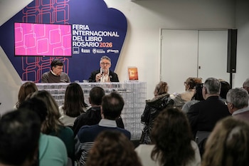 Dos hombres sentados detrás de un escritorio con un micrófono hablan ante un auditorio, frente a un cartel de la Feria del Libro de Rosario