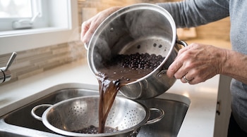 Manos de una persona vierten frijoles negros y agua caliente con vapor de una olla a un colador dentro de un fregadero de cocina.