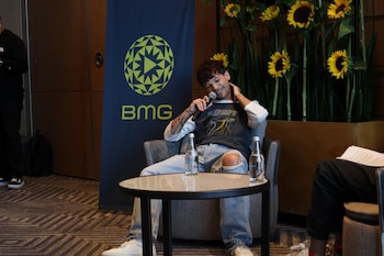 Hombre joven sentado en una silla gris, sonriendo y sosteniendo un micrófono. Viste camiseta oscura y jeans rotos. Detrás, banner azul de BMG y girasoles