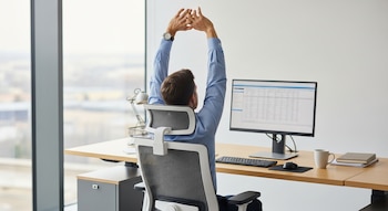 Un hombre en camisa azul se estira sentado en una silla de oficina frente a un escritorio con monitor, teclado y mouse junto a una ventana.