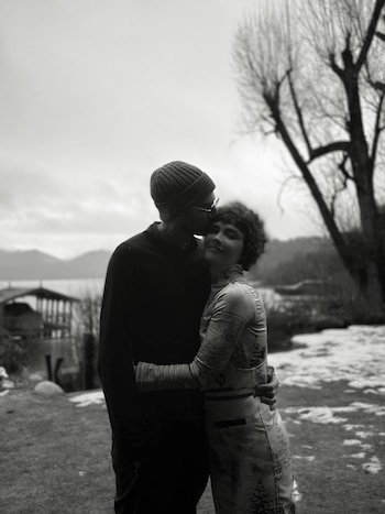 Fotografía en blanco y negro de una pareja abrazándose, con un hombre besando la frente de una mujer, junto a un lago y árboles secos con nieve