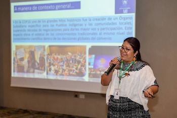 Una mujer con gafas y ropa tradicional sostiene un micrófono y un teléfono mientras habla frente a una pantalla de proyección con texto y fotos