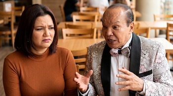 Monserrat Seminario con camiseta marrón y Melcochita con chaqueta floreada negra y rosa, gesticulando, sentados en un restaurante con mesas de madera.