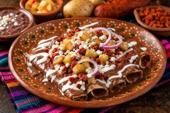 Un plato de barro con enfrijoladas cubiertas de frijol, chorizo, papa, queso, crema y cebolla morada, rodeado de recipientes con ingredientes.