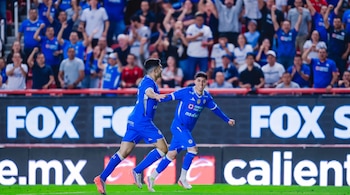 Dos jugadores de fútbol con uniforme azul corren en el campo mientras la afición en la tribuna celebra con los brazos en alto.