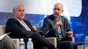 Dos hombres sentados en un panel. Uno a la derecha, de cabeza calva, con camisa azul y traje oscuro, sostiene un micrófono. El de la izquierda, de pelo gris, lleva traje oscuro y corbata rosa