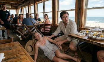 Mujer rubia en el suelo de un restaurante de madera, sostenida por un hombre. Mesas con mariscos crudos y comensales sorprendidos son visibles con el mar de fondo