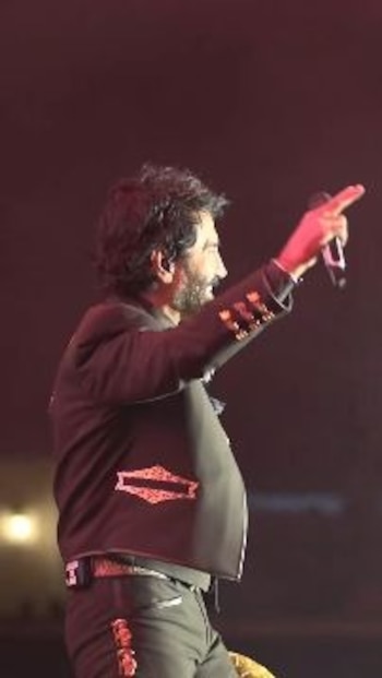 Hombre con cabello rizado y barba, vestido con traje de charro oscuro, sostiene un micrófono y señala con el dedo índice en un escenario iluminado