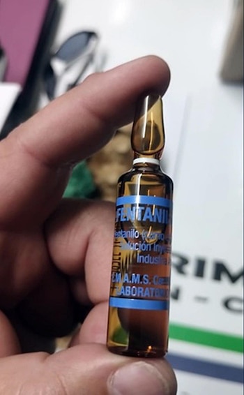 Primer plano de una mano sosteniendo una pequeña ampolla de cristal ámbar con una etiqueta azul que dice "FENTANIL" y otros textos en español