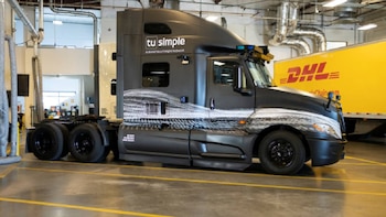 Un camión semi-remolque negro de TuSimple con detalles en blanco está estacionado en un almacén, junto a un remolque amarillo de DHL
