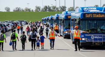 Estacionamiento exterior soleado con personas y autobuses azules LA Metro alineados con destino "SoFi Stadium". Empleados con chalecos naranjas guían a los asistentes.