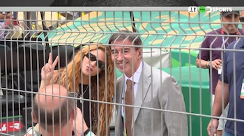 Shakira con gafas de sol y haciendo el signo de la paz, junto a un hombre sonriente en traje y corbata, ambos detrás de una valla metálica.
