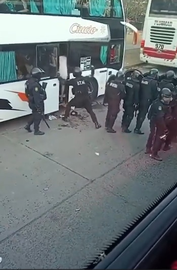Agentes de policía con cascos y escudos rodean la entrada de un autobús blanco de dos pisos estacionado en la calle