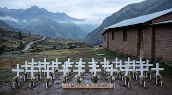 Veinticuatro cruces blancas con flores y velas alineadas frente a una casa de adobe, con montañas andinas y un letrero de madera en primer plano.