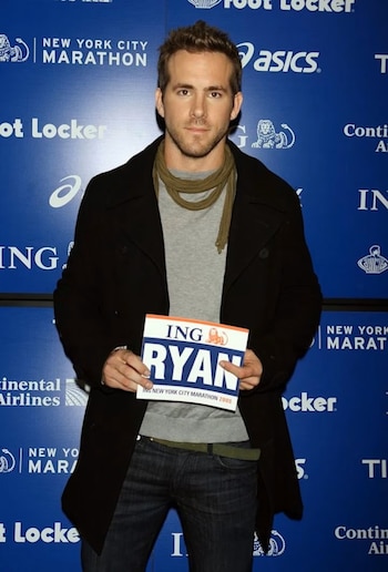 El actor Ryan Reynolds sostiene una cartela con su nombre frente a un panel publicitario de color azul con marcas patrocinadoras