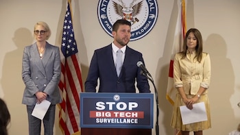 Un hombre habla en un podio junto a dos mujeres, situados entre dos banderas y frente al sello de la Fiscalía General del estado de Florida