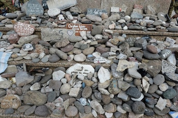 Montículo de piedras diversas, muchas con nombres y fechas escritas. Se observa una placa de madera y un trozo de tela con la bandera argentina