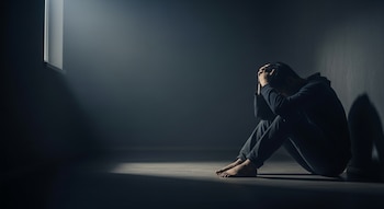 Una persona sentada en el suelo con la cabeza entre las manos, apoyada en una pared en un cuarto oscuro con una ventana.