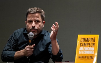 Martín Tetaz, un hombre barbudo con camisa oscura, sostiene un micrófono y gesticula con su mano derecha. A su derecha, un libro amarillo es visible